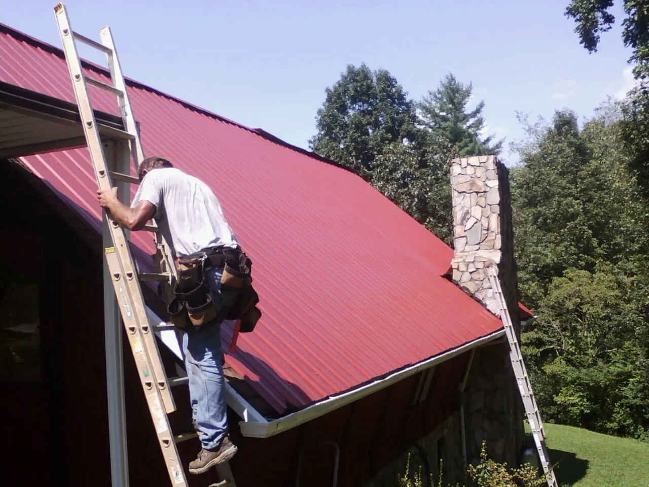Roofer on a ladder beside a red metal roof and stone chimney — Soffit & Fascia Repair by Roofing Brothers in Riverton